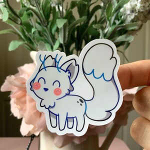 May include: A die-cut vinyl sticker featuring a white, fox-like creature with blue-tipped ears, a large bushy tail, and small blue antlers. The character has a smiling expression, pink circular cheeks, and two small black spots on its back.