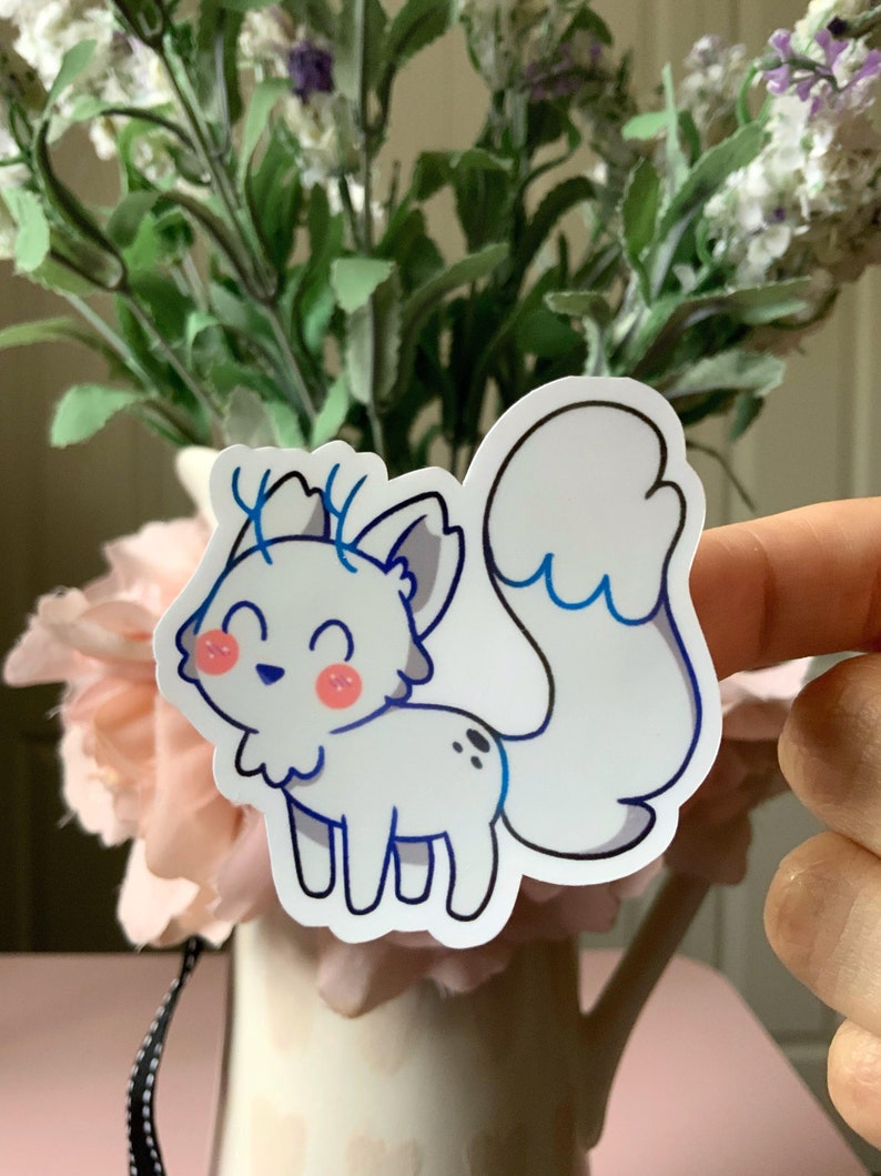 May include: A die-cut vinyl sticker featuring a white, fox-like creature with blue-tipped ears, a large bushy tail, and small blue antlers. The character has a smiling expression, pink circular cheeks, and two small black spots on its back.