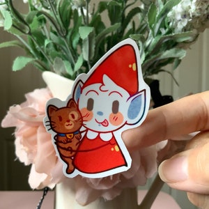 May include: A die-cut sticker featuring a cartoon character with pale blue skin, pointed ears, and a bright red pointed hat, wearing a red outfit with a white collar. The character is holding a small brown cat with a blue collar and yellow bell.