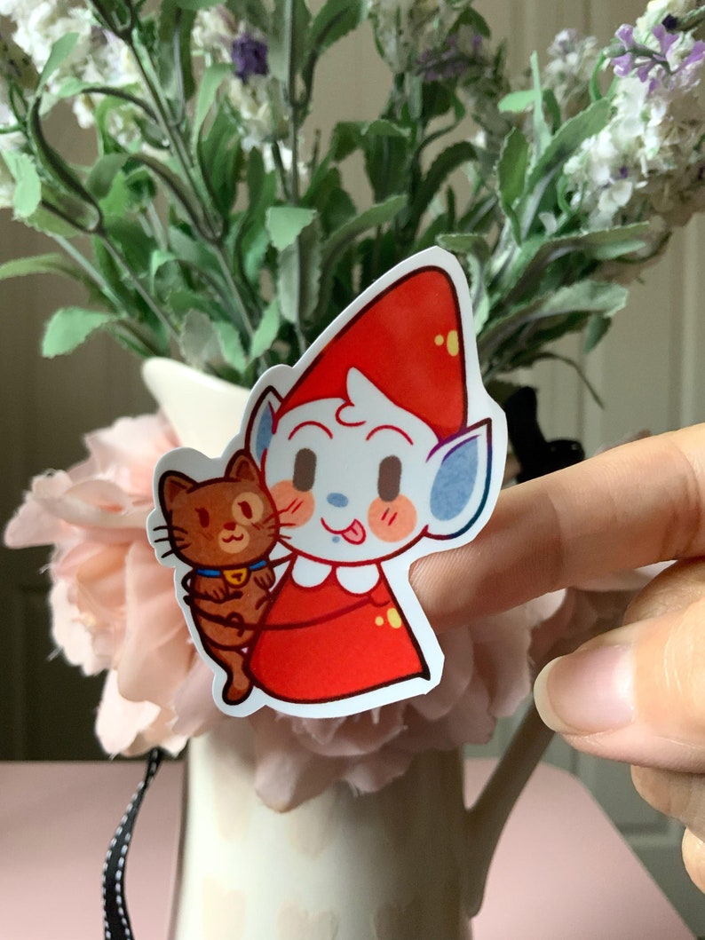 May include: A die-cut sticker featuring a cartoon character with pale blue skin, pointed ears, and a bright red pointed hat, wearing a red outfit with a white collar. The character is holding a small brown cat with a blue collar and yellow bell.