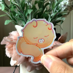 May include: A die-cut sticker featuring a stylized, sleeping orange creature with rounded ears and a curled tail. The illustration uses soft, warm gradients of yellow and orange with a thin, multi-colored outline, depicting a peaceful expression.