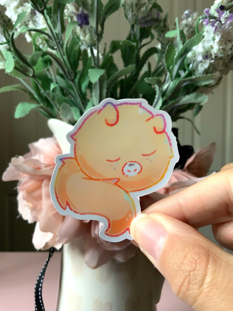 May include: A die-cut sticker featuring a stylized, sleeping orange creature with rounded ears and a curled tail. The illustration uses soft, warm gradients of yellow and orange with a thin, multi-colored outline, depicting a peaceful expression.