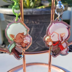 May include: Two clear acrylic keychains with colorful cartoon characters. The keychain on the left features a green-haired character holding a small, white character. The keychain on the right features a pink-haired character holding a small, white character.