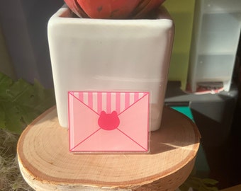May include: A pink envelope-shaped sticker featuring a cat face design. The sticker has a striped pattern on the top half and is placed on a wooden surface. A white square pot with a red plant is in the background.