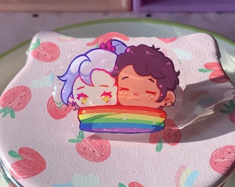 May include: A pink compact mirror with a rainbow design and a cartoon image of two people hugging. The mirror has a white background with pink strawberries.
