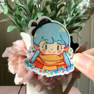 May include: A die-cut sticker featuring a cartoon character with light blue hair, a black hair bow, and a yellow scarf. The character is surrounded by small, colorful flowers in shades of blue, pink, and yellow against a white background.