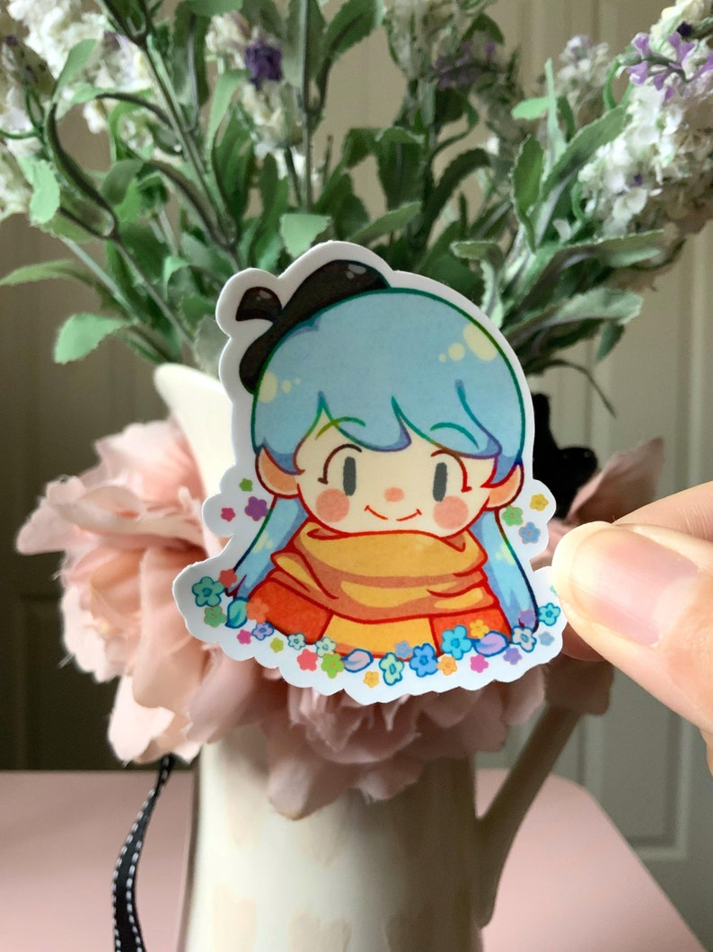 May include: A die-cut sticker featuring a cartoon character with light blue hair, a black hair bow, and a yellow scarf. The character is surrounded by small, colorful flowers in shades of blue, pink, and yellow against a white background.