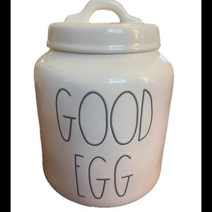 Puede incluir: Tarro de cerámica blanca con tapa y asa, con las palabras "GOOD EGG" en letras grises. El tarro es un recipiente de almacenamiento de cocina, probablemente para huevos u otros artículos pequeños.