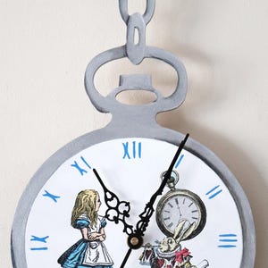 May include: A silver pocket watch clock with a white face and blue Roman numerals. The clock face features Alice in Wonderland characters, including Alice, the White Rabbit, and a pie.