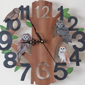 May include: A wooden clock shaped like a tree trunk with black numbers and owl illustrations. The owls are painted in realistic colours and are perched on the branches of the tree. The clock face is brown with black hands.