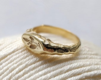 Può includere: Un anello d'oro con un design testurizzato e organico. L'anello ha una parte superiore ovale in rilievo con un motivo dettagliato.