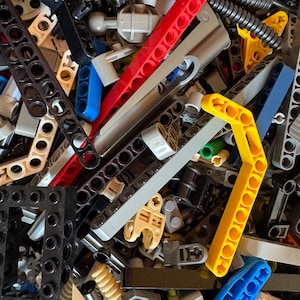 May include: A close-up view of a pile of colorful plastic building blocks. The blocks are in various shapes and sizes, including long beams, angled pieces, and connectors. Colors include red, yellow, blue, black, gray, and tan. The pieces are scattered.