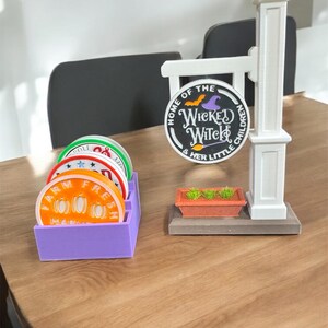 Puede incluir: Una decoración temática de Halloween con un letrero que dice "Home of the Wicked Witch & Her Little Children". El conjunto incluye una bandeja morada con posavasos redondos y una pequeña jardinera. Los posavasos tienen varios diseños.