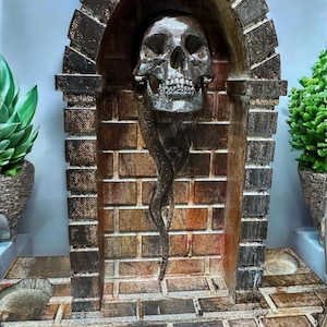 May include: A decorative sculpture featuring a metallic skull within a brick archway. The skull has detailed features and is suspended above a curved, elongated element. The brickwork is in shades of brown and orange, creating a gothic or Halloween theme.