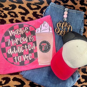 May include: A hot pink t-shirt with the text "Maybe 7 Brew is addicted to me" and a graphic of a coffee drink. A red and white baseball cap, blue jeans, and a wooden monogram keychain are also in the image.