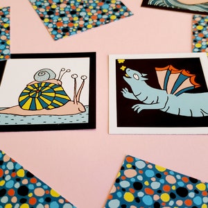 Peut inclure: Deux impressions d'art carr&eacute;es avec des illustrations fantaisistes. L'une montre un escargot avec une coquille jaune et bleue, l'autre un dragon bleu avec des ailes orange et bleues. Des cartes &agrave; motifs de points color&eacute;s sont &eacute;parpill&eacute;es.