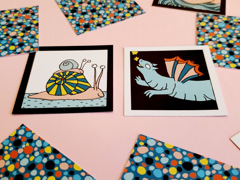 Peut inclure: Deux impressions d'art carr&eacute;es avec des illustrations fantaisistes. L'une montre un escargot avec une coquille jaune et bleue, l'autre un dragon bleu avec des ailes orange et bleues. Des cartes &agrave; motifs de points color&eacute;s sont &eacute;parpill&eacute;es.