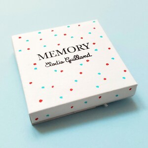 Peut inclure: Une bo&icirc;te blanche orn&eacute;e de pois rouges et bleus. Le mot "MEMORY" est imprim&eacute; en noir, avec "Elodie Guillaud" en dessous dans une police script. La bo&icirc;te est pos&eacute;e sur un fond bleu clair.