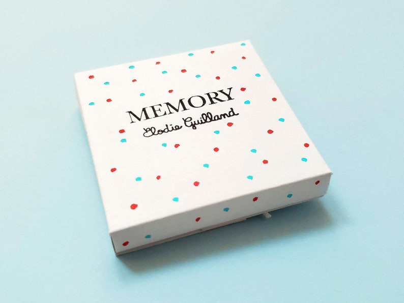 Peut inclure: Une bo&icirc;te blanche orn&eacute;e de pois rouges et bleus. Le mot "MEMORY" est imprim&eacute; en noir, avec "Elodie Guillaud" en dessous dans une police script. La bo&icirc;te est pos&eacute;e sur un fond bleu clair.