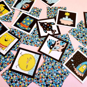 Peut inclure: Un ensemble de cartes illustr&eacute;es avec des bordures noires, repr&eacute;sentant des personnages et des objets fantaisistes. Les cartes sont dispers&eacute;es sur une surface rose, entrecoup&eacute;es de carr&eacute;s de papier &agrave; motifs bleus, oranges et jaunes. Les illustrations incluent un chien, une glace et une fus&eacute;e.