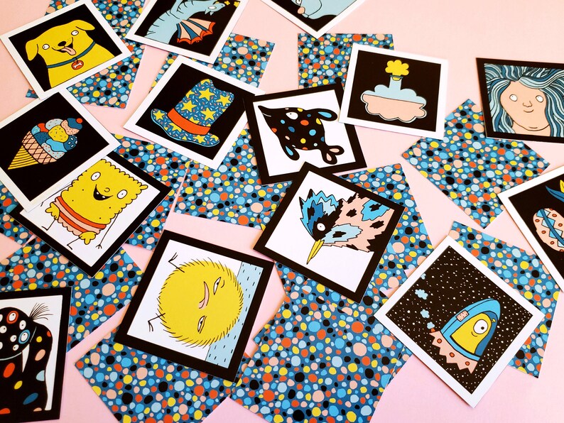 Peut inclure: Un ensemble de cartes illustr&eacute;es avec des bordures noires, repr&eacute;sentant des personnages et des objets fantaisistes. Les cartes sont dispers&eacute;es sur une surface rose, entrecoup&eacute;es de carr&eacute;s de papier &agrave; motifs bleus, oranges et jaunes. Les illustrations incluent un chien, une glace et une fus&eacute;e.