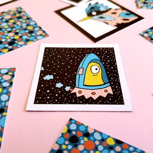 Peut inclure: Une impression d'art carr&eacute;e repr&eacute;sentant un extraterrestre de dessin anim&eacute; dans un vaisseau spatial bleu et jaune sur un fond noir &eacute;toil&eacute;. L'impression est entour&eacute;e d'autres impressions avec des motifs de points color&eacute;s, le tout sur une surface rose.