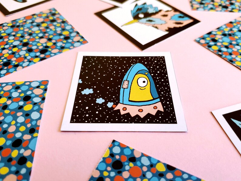 Peut inclure: Une impression d'art carr&eacute;e repr&eacute;sentant un extraterrestre de dessin anim&eacute; dans un vaisseau spatial bleu et jaune sur un fond noir &eacute;toil&eacute;. L'impression est entour&eacute;e d'autres impressions avec des motifs de points color&eacute;s, le tout sur une surface rose.