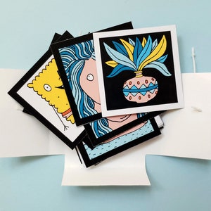 Peut inclure: Ensemble d'impressions d'art carr&eacute;es avec des bordures noires, pr&eacute;sentant des illustrations fantaisistes. Une impression montre un personnage jaune de dessin anim&eacute;, une autre une femme aux cheveux bleus et une troisi&egrave;me une plante dans un pot d&eacute;coratif. Les impressions sont pr&eacute;sent&eacute;es dans une bo&icirc;te blanche.