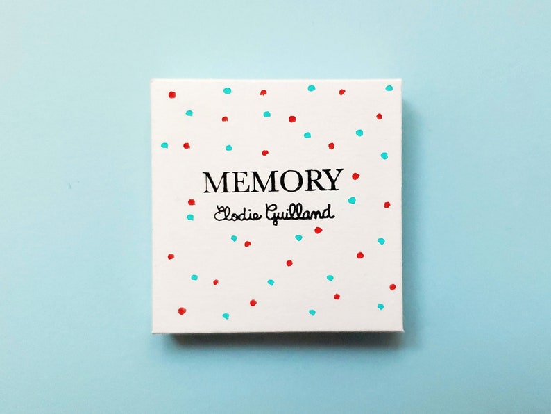 Peut inclure: Une bo&icirc;te carr&eacute;e blanche avec le mot &laquo;&nbsp;MEMORY&nbsp;&raquo; en noir et le nom &laquo;&nbsp;Elodie Guilland&nbsp;&raquo; en cursive. La bo&icirc;te est d&eacute;cor&eacute;e de points rouges et turquoise &eacute;parpill&eacute;s. Le fond est bleu clair uni.