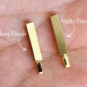 May include: Two gold-coloured rectangular bar pendants. One pendant features a reflective, polished shiny finish, while the other pendant displays a textured, brushed matte finish. Each bar includes a small attached bail at the bottom for hanging.