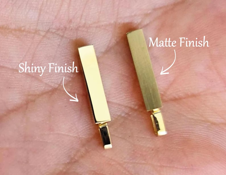 May include: Two gold-coloured rectangular bar pendants. One pendant features a reflective, polished shiny finish, while the other pendant displays a textured, brushed matte finish. Each bar includes a small attached bail at the bottom for hanging.