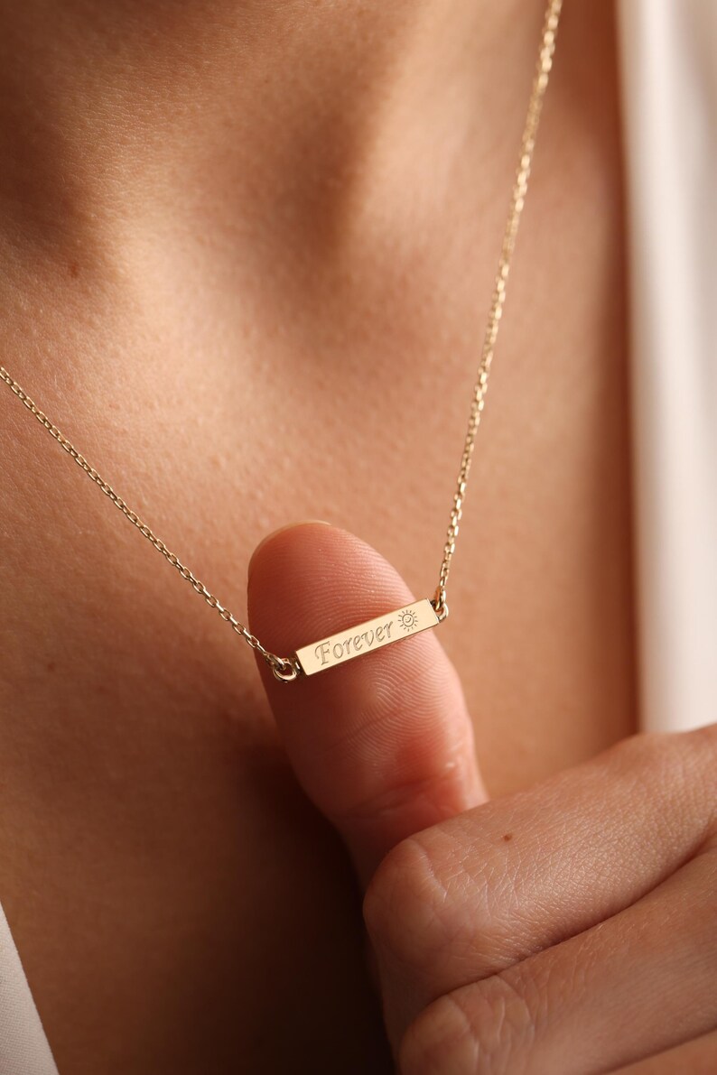 May include: Gold necklace with a small bar pendant that says "Forever" with a sun symbol.