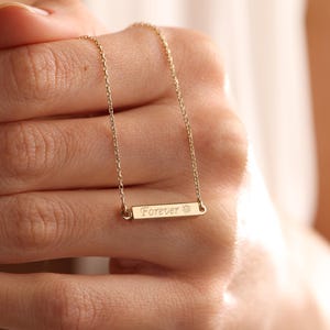 May include: A gold necklace with a thin chain and a small bar pendant that says "Forever" with a sun symbol.