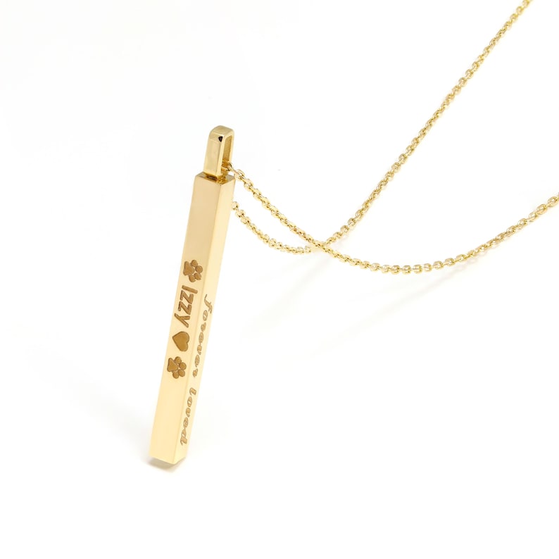 May include: A gold-toned vertical bar pendant necklace featuring an engraved design with the name Izzy, a heart symbol, two paw prints, and the text forever loved. The pendant hangs from a delicate matching gold-toned chain link necklace.