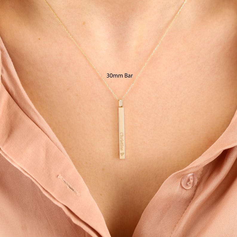 May include: A gold-toned necklace featuring a vertical bar pendant measuring 30mm in length. The bar is engraved with the name Christine followed by a small heart symbol. The pendant hangs from a delicate chain resting against a light-coloured fabric.