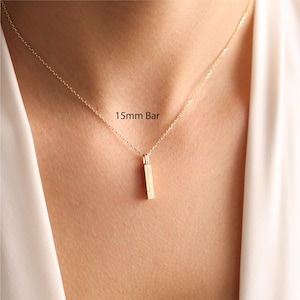 May include: A gold-toned necklace featuring a delicate chain and a vertical 15mm bar pendant. The pendant is engraved with text and hangs centrally from the chain. The piece is worn against the skin, showcasing the metallic finish and design.