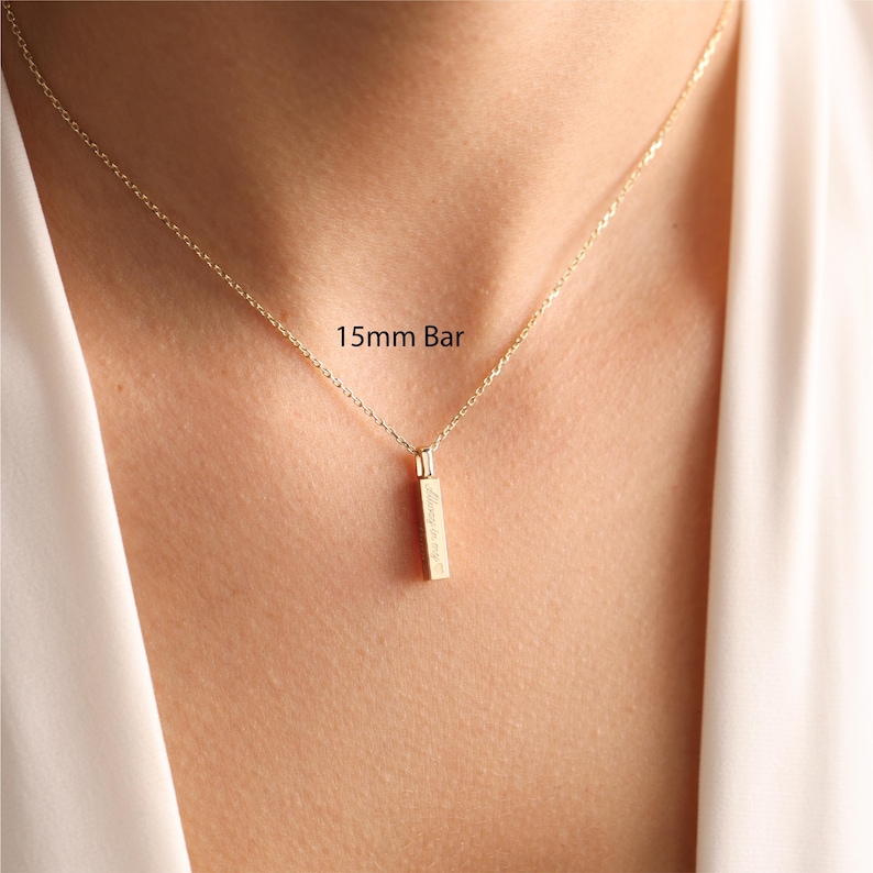 May include: A gold-toned necklace featuring a delicate chain and a vertical 15mm bar pendant. The pendant is engraved with text and hangs centrally from the chain. The piece is worn against the skin, showcasing the metallic finish and design.
