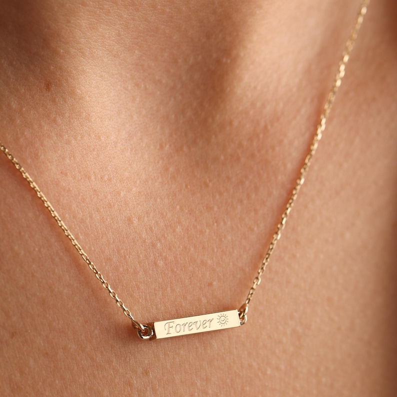 May include: A gold chain necklace with a small bar pendant that says "Forever" with a sun symbol.