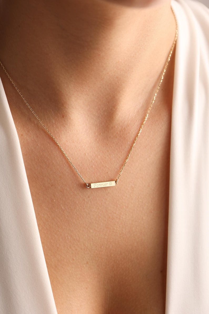 May include: A gold necklace with a delicate chain and a small bar pendant that says "Forever" with a tiny diamond accent.