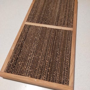 Könnte beinhalten: Rechteckiges Kratzbrett für Katzen mit einem Holzrahmen. Die Kratzfläche besteht aus Wellpappe und bietet eine strukturierte Fläche zum Kratzen für Katzen. Der Rahmen ist aus hellem Holz gefertigt.