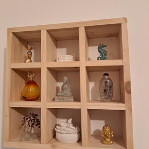 Puede incluir: Una estanter&iacute;a de madera con nueve compartimentos cuadrados, cada uno con un objeto decorativo diferente. Los objetos incluyen una figurita de gato dorada, una caja de cer&aacute;mica blanca, un caballito de mar azul, una botella de vidrio con tapa dorada, una peque&ntilde;a estatua de Buda gris, una botella de vidrio transparente con etiqueta, un peque&ntilde;o &aacute;rbol con ramas marrones, una figurita de &aacute;ngel de cer&aacute;mica blanca y una figurita dorada de una persona con cabeza grande.