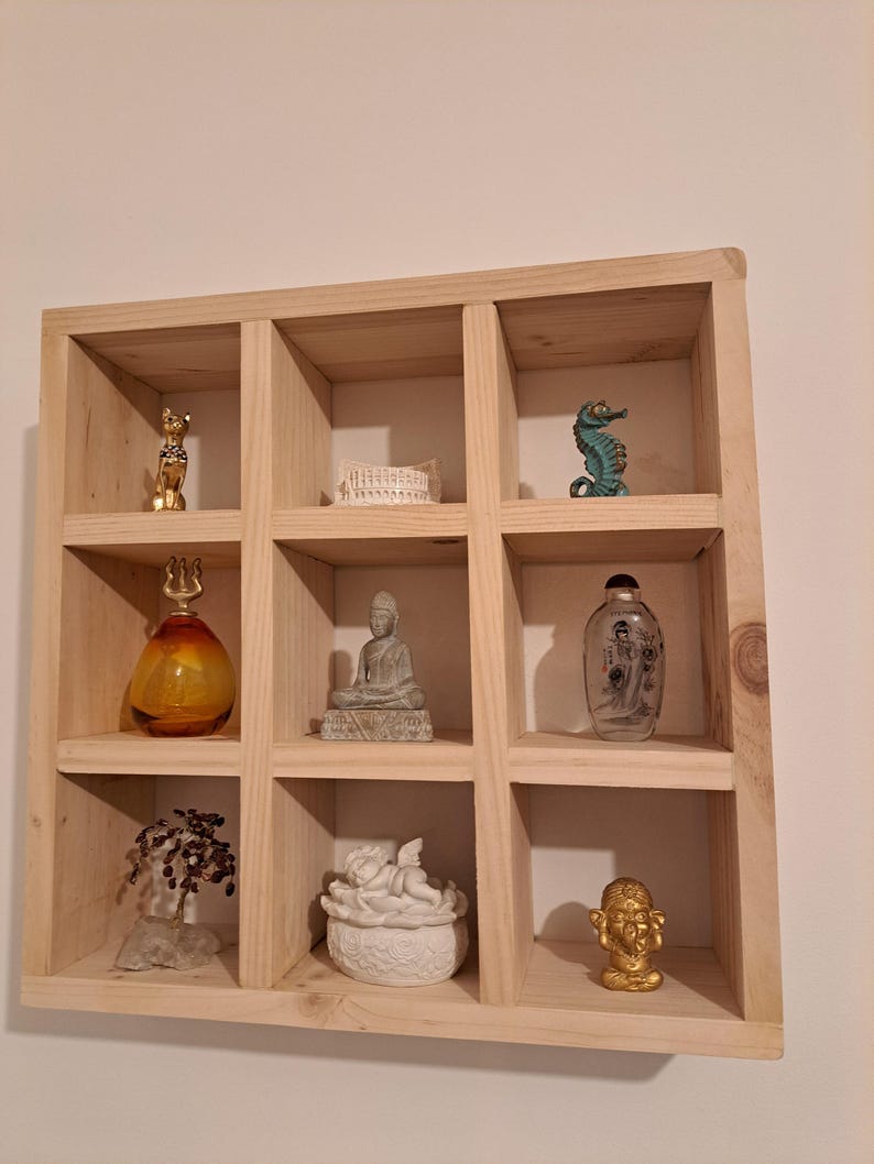 Puede incluir: Una estanter&iacute;a de madera con nueve compartimentos cuadrados, cada uno con un objeto decorativo diferente. Los objetos incluyen una figurita de gato dorada, una caja de cer&aacute;mica blanca, un caballito de mar azul, una botella de vidrio con tapa dorada, una peque&ntilde;a estatua de Buda gris, una botella de vidrio transparente con etiqueta, un peque&ntilde;o &aacute;rbol con ramas marrones, una figurita de &aacute;ngel de cer&aacute;mica blanca y una figurita dorada de una persona con cabeza grande.