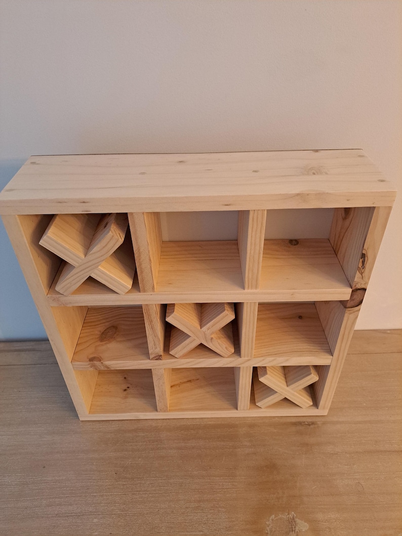 Puede incluir: Un juego de tres en raya de madera con nueve compartimentos. El juego est&aacute; hecho de madera sin terminar y tiene tres piezas en forma de X en su lugar.
