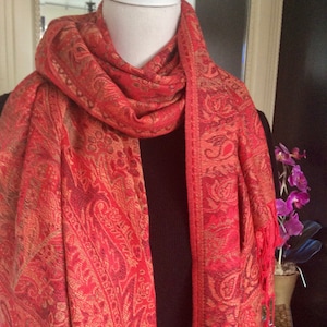 May include: A red and gold paisley patterned scarf with fringe. The scarf is draped over a mannequin head and shoulders. The scarf has a detailed design and a warm colour palette.