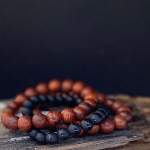 May include: Two beaded bracelets. One bracelet features dark brown wooden beads, and the other has black matte beads. The bracelets are resting on a piece of weathered wood against a dark background.