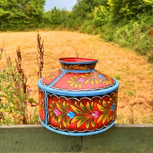 May include: A hand-painted, round container with a red lid and body, decorated with floral patterns in yellow, pink, and blue against a black background. It has a blue rim and base. The background shows a field and trees.