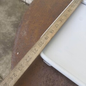 Puede incluir: Una regla de madera con marcas negras, que mide hasta 61 cm, se encuentra en diagonal sobre una superficie blanca y una superficie de metal marr&oacute;n oxidado. La regla est&aacute; en &aacute;ngulo, mostrando los n&uacute;meros del 12 al 24.