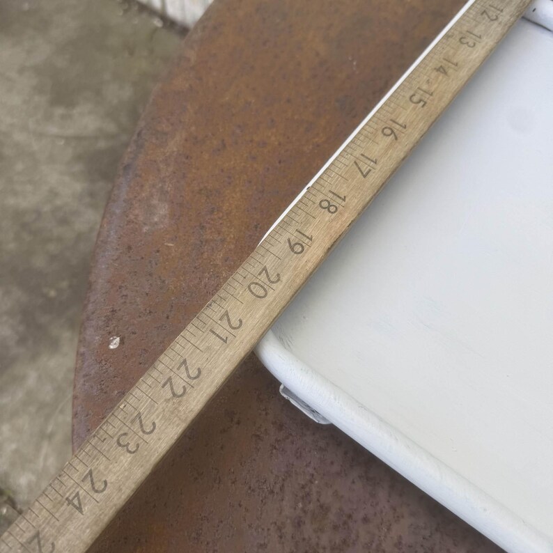 Puede incluir: Una regla de madera con marcas negras, que mide hasta 61 cm, se encuentra en diagonal sobre una superficie blanca y una superficie de metal marr&oacute;n oxidado. La regla est&aacute; en &aacute;ngulo, mostrando los n&uacute;meros del 12 al 24.