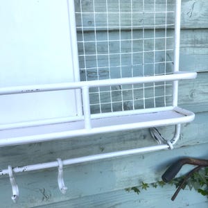 Puede incluir: Organizador de pared de metal blanco con un estante, ganchos y un panel de rejilla de alambre. El organizador tiene forma rectangular y est&aacute; dise&ntilde;ado para almacenamiento y exhibici&oacute;n. Los ganchos son curvos y el estante es plano.