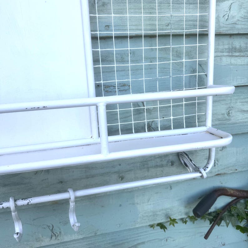 Puede incluir: Organizador de pared de metal blanco con un estante, ganchos y un panel de rejilla de alambre. El organizador tiene forma rectangular y est&aacute; dise&ntilde;ado para almacenamiento y exhibici&oacute;n. Los ganchos son curvos y el estante es plano.